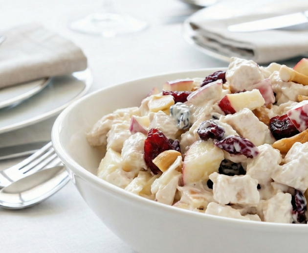 O salată bogată de pui cu iaurt grecesc, cu merișoare roșii, felii de măr și nuci, servită într-un bol alb adânc pe o masă elegantă cu decor minimalist.