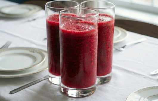 Trei pahare înalte, cilindrice, pline cu un smoothie dens de culoare roșu-purpuriu intens, așezate pe o masă cu față de masă albă și veselă elegantă.