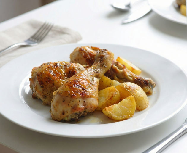 O porție apetisantă de copănele de pui la cuptor cu piele crocantă și condimentată, servite alături de cartofi rumeniți aurii pe o farfurie albă, într-un cadru luminos de masă.