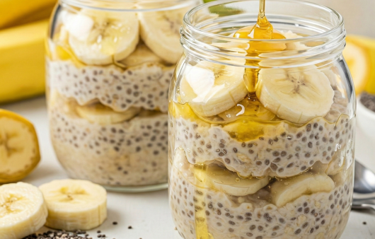 Două borcane de sticlă pline cu budincă de chia și ovăz, stratificate cu felii de banană și miere lichidă, pe o masă albă cu semințe de chia presărate.