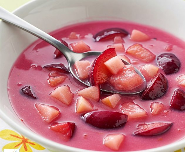 Un bol alb plin cu supă de prune de un roz intens, conținând felii de prune proaspete și cubulețe de fructe, cu o lingură metalică ce ridică o porție generoasă, așezat pe o față de masă cu motive florale colorate.