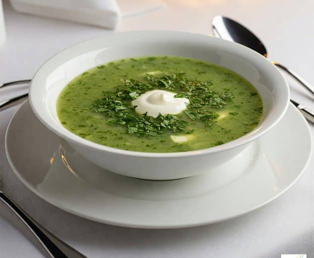 Un bol alb de ceramică umplut cu supă de pătrunjel verde, decorată cu mult pătrunjel proaspăt tocat și o porție centrală de smântână cremoasă, servită pe o farfurie cu o lingură argintie pe o față de masă albă.