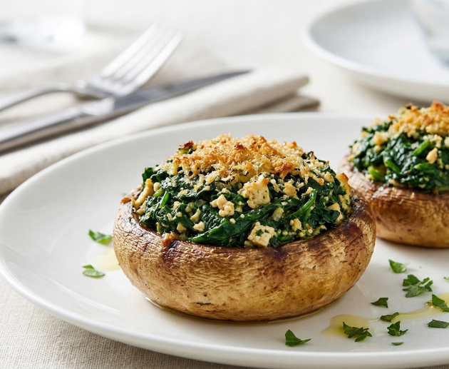 Două ciuperci mari de tip Champignon, umplute cu un amestec de spanac și tofu, acoperite cu drojdie de bere auriu, servite pe o farfurie albă elegantă.