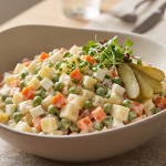 Un bol plin cu salată franțuzească cremoasă, preparată din legume fierte tăiate cubulețe, decorat cu arpagic tocat fin, microplante și felii de castravete murat, așezat pe o masă elegantă.