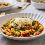 O farfurie albă cu margini ondulate conținând paste fusilli cu sos de roșii, frunze de spanac și un strat generos de cașcaval ras, așezată pe o față de masă gri lângă un pahar cu apă.