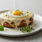 Un sandviș Croque Madame pe o farfurie albă, acoperit cu un strat generos de sos béchamel care curge pe margini, cu un ou prăjit tip ochi în centru, garnisit cu pătrunjel proaspăt tocat mărunt și microplante, alături de tacâmuri argintii, pe o față de masă din in gri.
