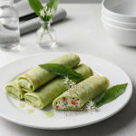 O farfurie albă cu patru clătite verzi rulate, dintre care una secționată arătând o umplutură cremoasă cu ardei, decorate cu frunze și flori proaspete de leurdă, alături de tacâmuri negre pe un fundal gri deschis.