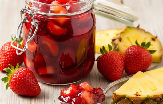 Un borcan de sticlă cu gem de căpșuni și ananas, înconjurat de căpșuni proaspete și felii de ananas pe o masă din lemn deschis la culoare, cu o lingură plină de gem în prim-plan.