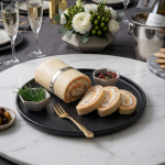 Un platou negru elegant cu o ruladă de brânză umplută cu șuncă și verdețuri, feliată parțial, așezată pe un platou de marmură albă, într-un decor festiv cu pahare de șampanie, flori și biscuiți sărați.