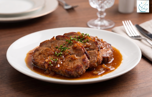 O farfurie albă din porțelan cu ceafă de porc la cuptor, acoperită cu ceapă caramelizată și sos de bere brună, garnisit cu arpagic verde tocat, așezată pe o masă de lemn alături de o furculiță și un pahar de cristal.