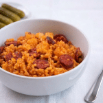 Un bol alb plin cu cuș-cuș gătit cu bucăți de cartofi și felii de cârnați, servit pe o față de masă albă alături de o porție de castraveți murați și o furculiță.