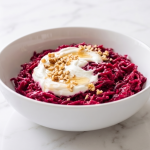 Un bol alb cu salată de sfeclă roșie răzuită, acoperită cu un strat de iaurt cremos, miere și nuci mărunțite, așezat pe o suprafață de marmură.