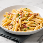 O farfurie adâncă albă plină cu paste penne acoperite cu un sos cremos de brânză, bucăți de bacon crocant și pătrunjel proaspăt, așezată pe un șervet gri pe un blat de marmură albă.