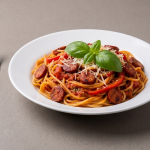 O farfurie albă cu spaghete amestecate cu sos de roșii, rondele de cârnați rumeniți și fâșii de ardei kapia roșu, presărate cu brânză rasă și decorate cu frunze de busuioc, așezată pe un blat gri mat, alături de o furculiță.