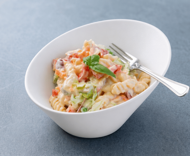 Un bol alb asimetric conținând o porție de salată de paste fusilli cremoasă cu legume cubulețe, o furculiță de argint și o garnitură centrală de busuioc, pe un fundal gri texturat.