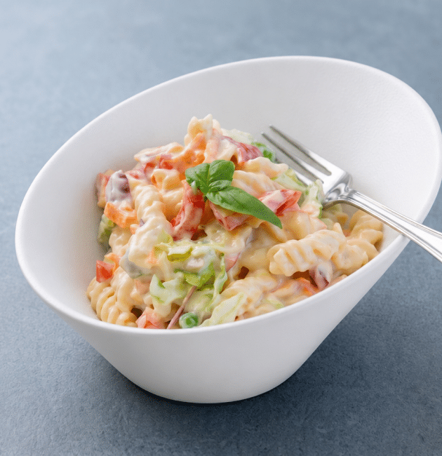 Un bol alb asimetric conținând o porție de salată de paste fusilli cremoasă cu legume cubulețe, o furculiță de argint și o garnitură centrală de busuioc, pe un fundal gri texturat.