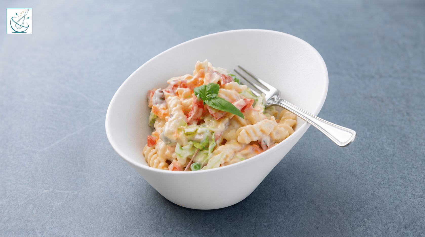 Un bol alb asimetric conținând o porție de salată de paste fusilli cremoasă cu legume cubulețe, o furculiță de argint și o garnitură centrală de busuioc, pe un fundal gri texturat.
