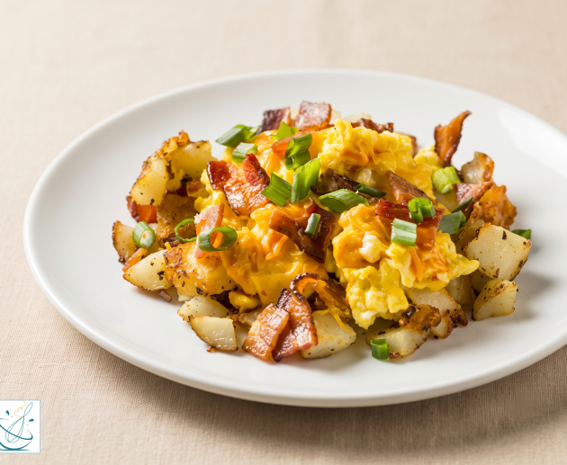 O farfurie albă plată plin cu cartofi prăjiți cubulețe, ouă omletă cremoase și bucăți crocante de bacon, garnisit cu ceapă verde tocată, servit pe o suprafață texturată bej.