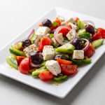 Un platou alb dreptunghiular plin cu o salată proaspătă din roșii, castraveți, ceapă roșie, ardei verde, măsline Kalamata și cuburi mari de brânză feta presărate cu oregano, pe un fundal gri deschis.