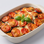 Un vas termorezistent din ceramică plin cu vinete și roșii feliate, gratinate la cuptor cu o crustă aurie de brânză, decorate central cu frunze proaspete de busuioc, pe un fundal alb-cenușiu.