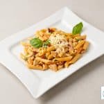 O farfurie albă dreptunghiulară cu paste penne, bucăți de pui și roșii uscate, presărate cu brânză rasă și decorate cu frunze de busuioc proaspăt, pe un fundal bej deschis.