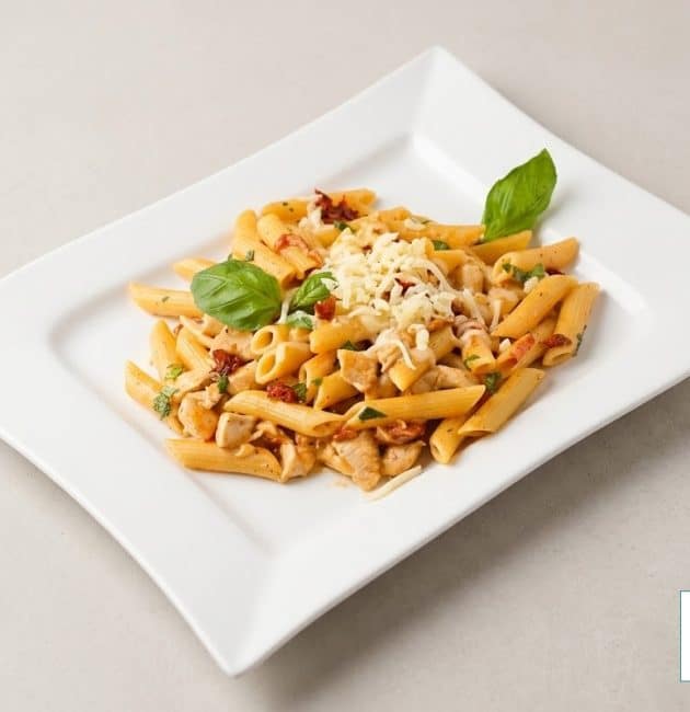 O farfurie albă dreptunghiulară cu paste penne, bucăți de pui și roșii uscate, presărate cu brânză rasă și decorate cu frunze de busuioc proaspăt, pe un fundal bej deschis.
