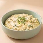 Un bol rotund de culoare verde pastel plin cu cremă de ouă texturată, garnisit central cu mărar și cepșoară proaspătă, pe un fundal bej-piersică scăldat în lumină naturală.