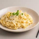 O porție generoasă de paste Fettuccine Alfredo cu sos cremos, parmezan ras, piper negru și frunze proaspete de verdeață, servite într-o farfurie din ceramică, alături de o furculiță neagră mată.