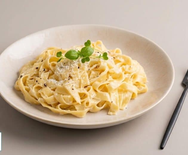 O porție generoasă de paste Fettuccine Alfredo cu sos cremos, parmezan ras, piper negru și frunze proaspete de verdeață, servite într-o farfurie din ceramică, alături de o furculiță neagră mată.